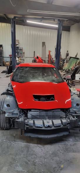 Red Corvette with the front bumper off, parked between two lift posts inside the shop